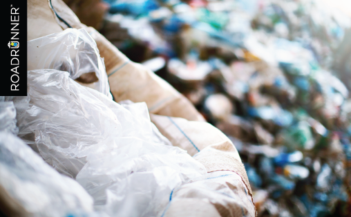 Plastic bags inside a cardboard box with a mound of more assorted recyclables in the background.